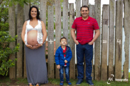 gestante, filho e marido na frente de uma cerca de madeira onde se vê o sinal de mais somando os três e resultando em quatro